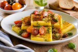 Kalte Polenta-Schnitten mit Tomaten auf einem weißen Teller, garniert mit frischem Basilikum und Olivenöl, Sommerrezept